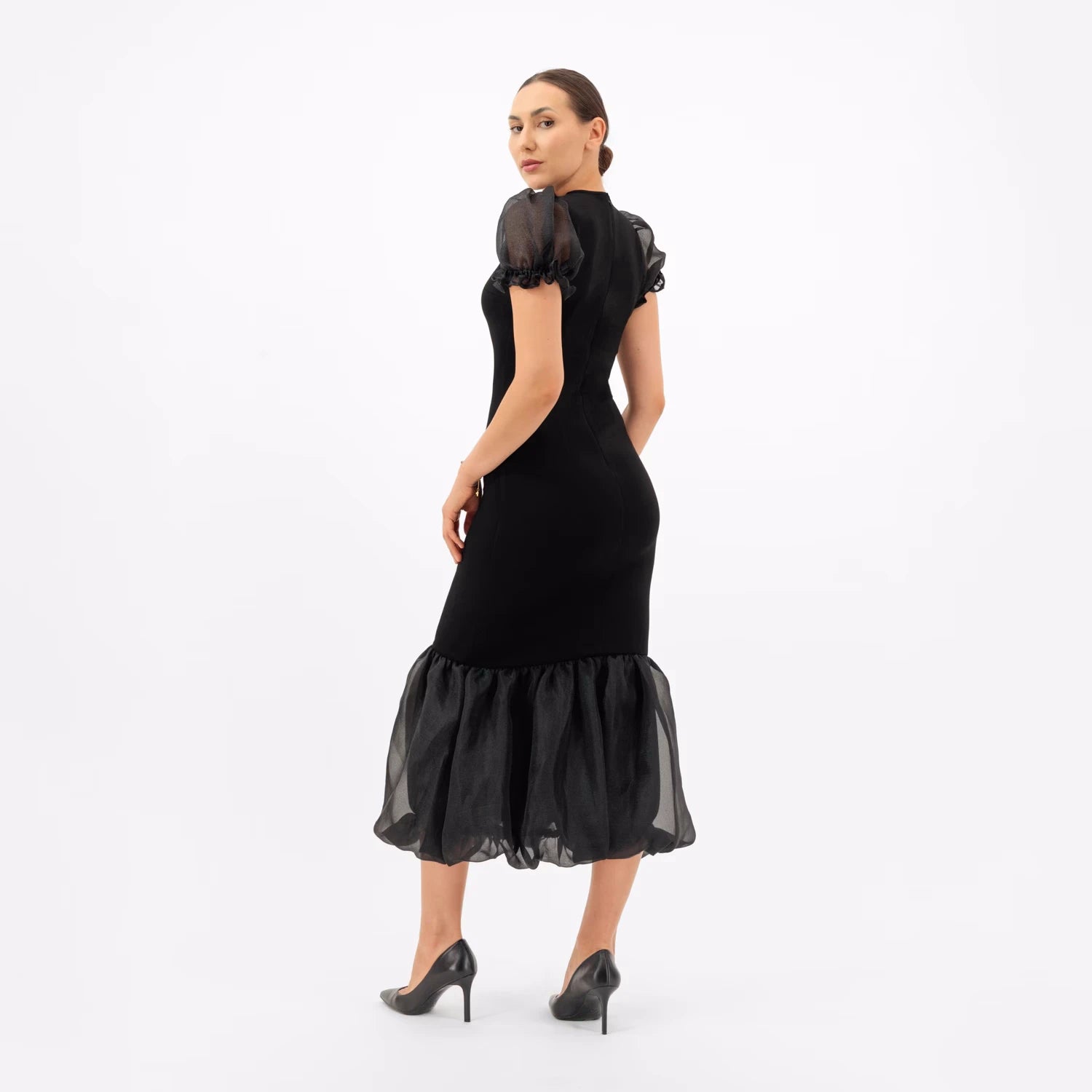 A woman from a three-quarter back view, wearing a obsidian black maxi dress, puffed sleeves and a bubble hem.